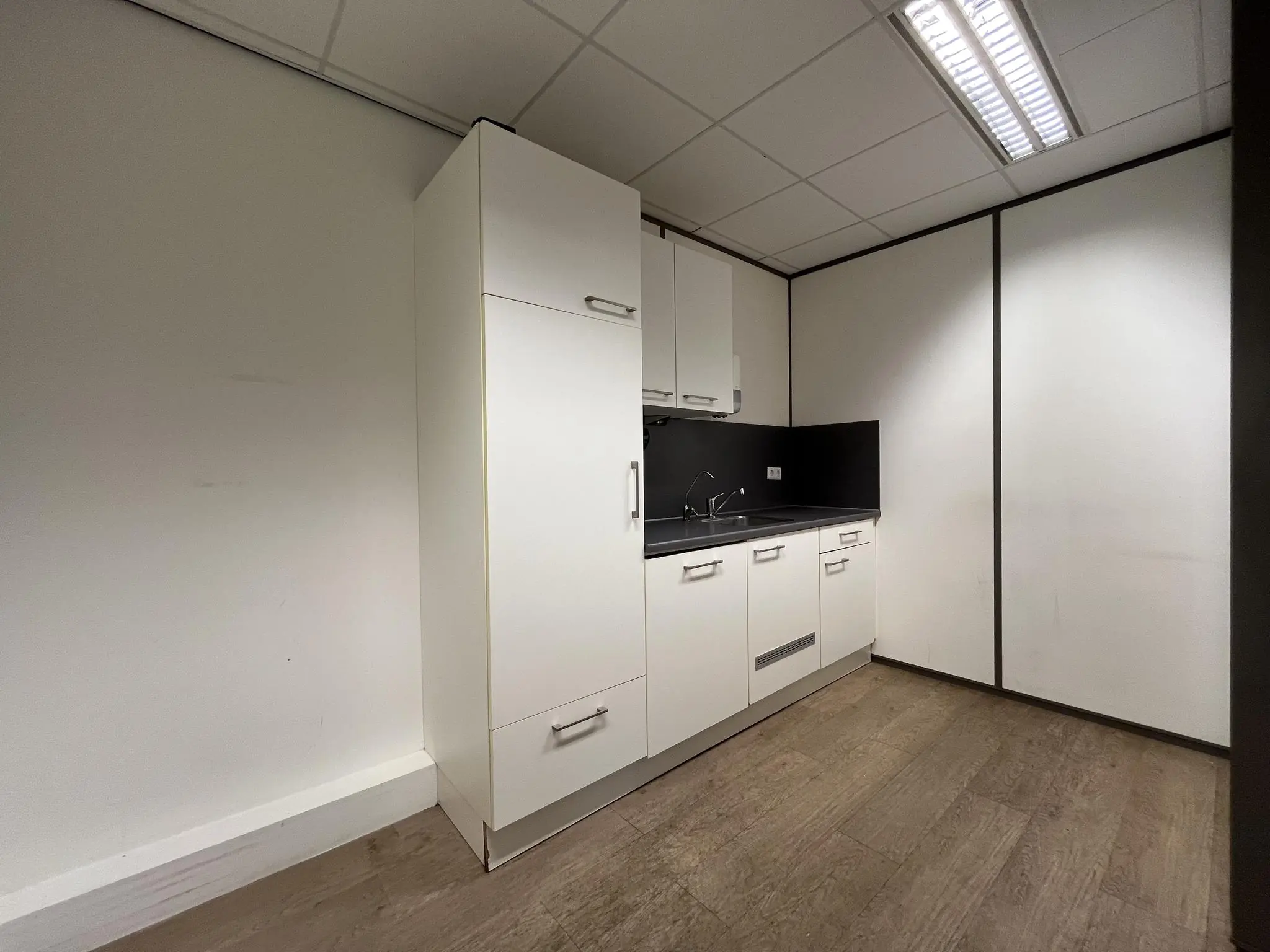 Kleine pantry met witte keukenkastjes en zwart werkblad in een kantoorpand aan de Burgemeester Verderlaan.